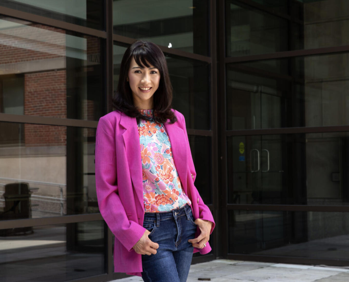 Woman posing in front of large windows wearing a pink suit jacket, floral shirt and jeans.