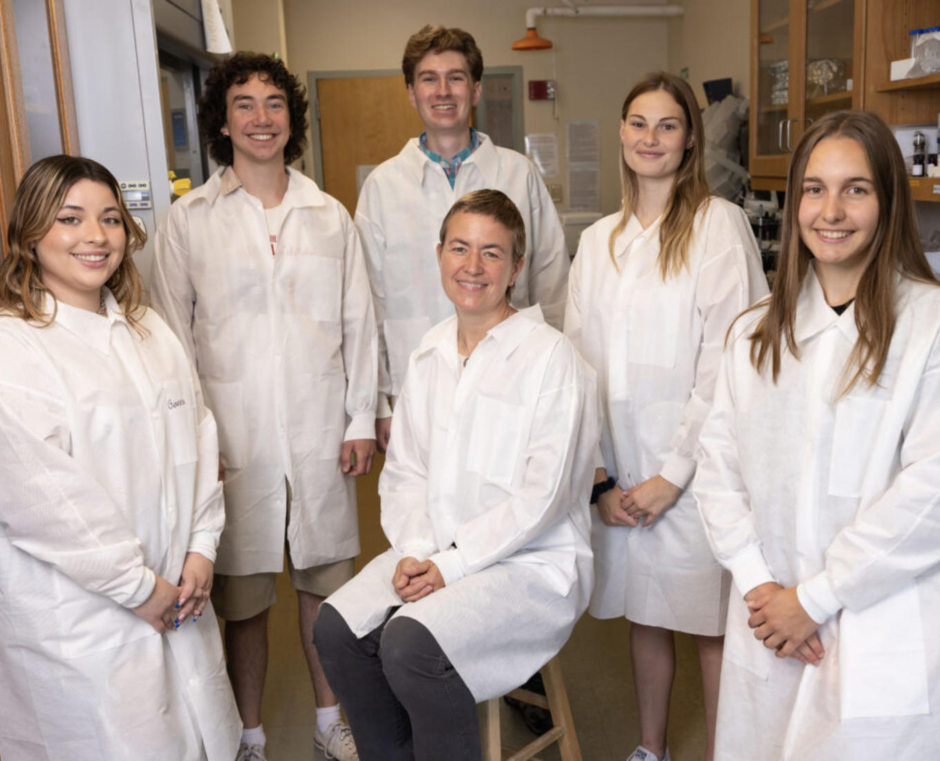 Biology students dressed in white lab coats stand behind their professor, who is seated in the center.