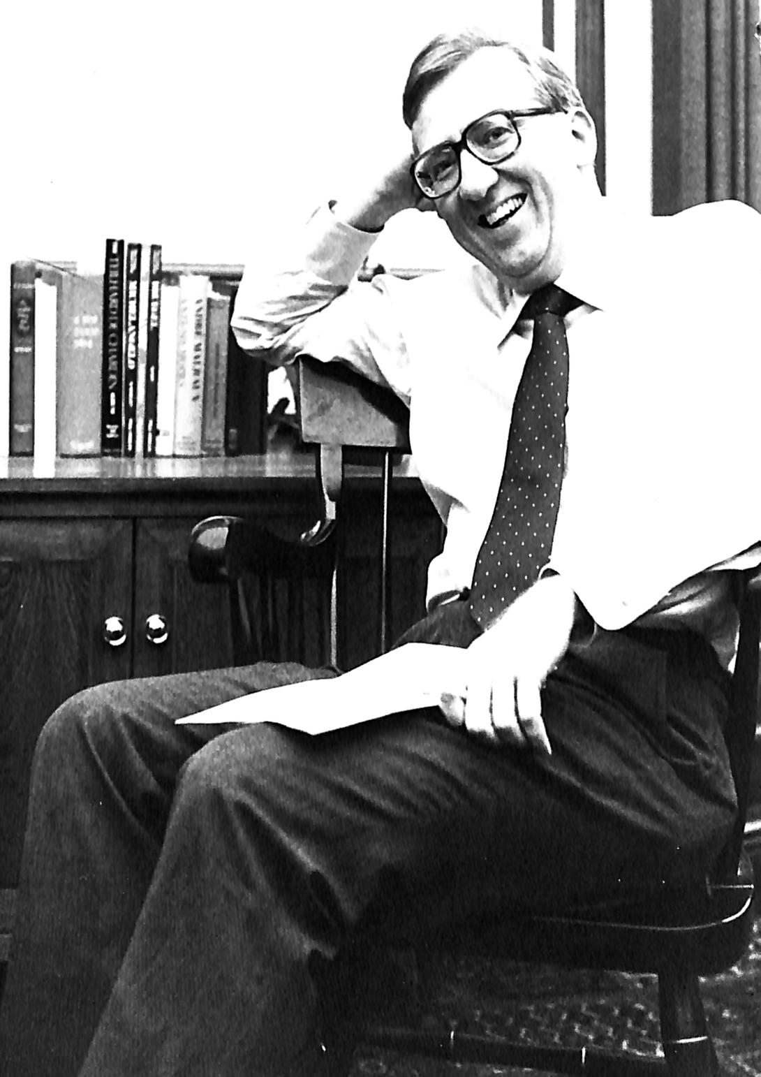 Black and white photo of a middle-aged man with glasses in a white shirt, dark tie sitting and leaning on a desk