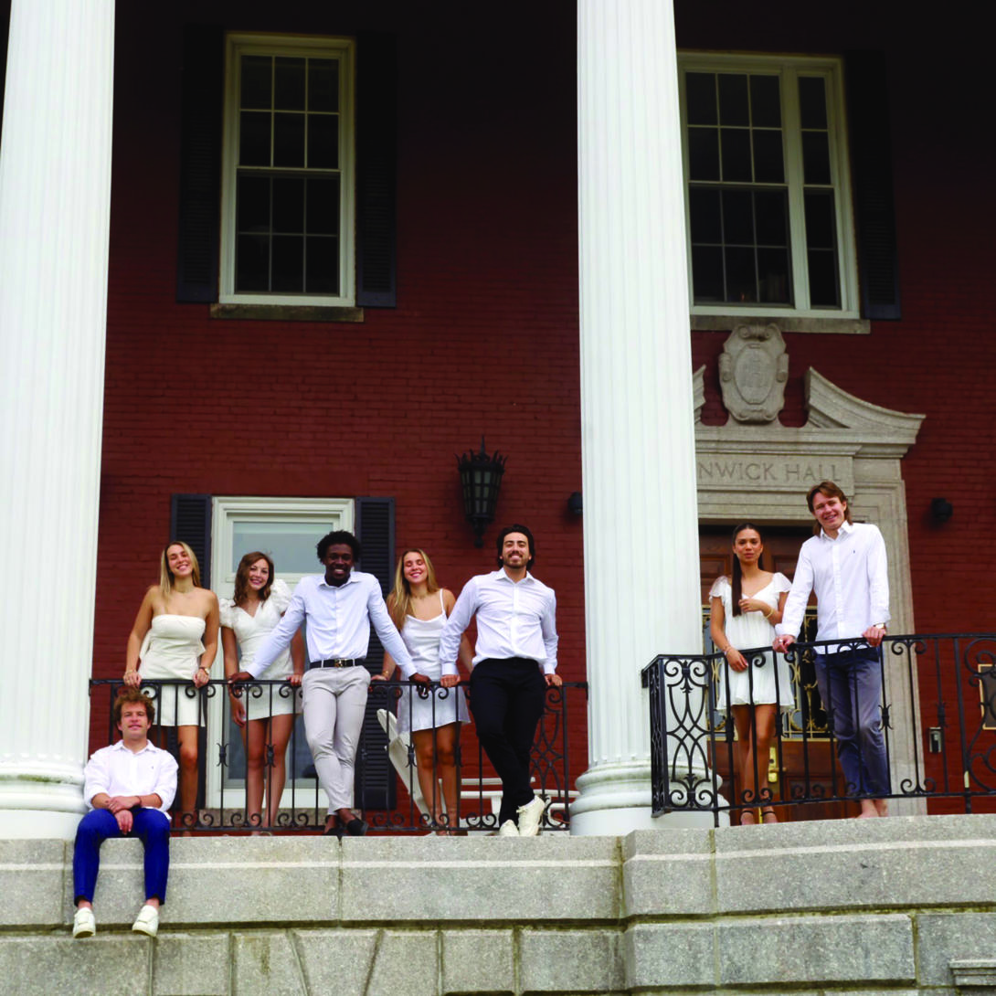 A group of students standing on a porch next to a pillar and a brick building 
